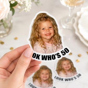 Op de afbeelding: Een gestanste sticker met een foto van een jong kind met krullend blond haar in een lichtgekleurde top. Onder de afbeelding van het kind staat de tekst "LOOK WHO'S 50" gedrukt in vette, zwarte, schreefloze hoofdletters op een witte achtergrond.