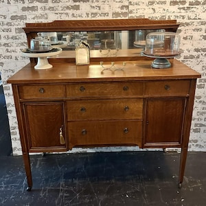 May include: A wooden buffet with a mirror, drawers, and cabinet doors. The buffet is a warm brown color and has a rectangular shape. There are decorative items on top, including a cake stand and a small framed picture.