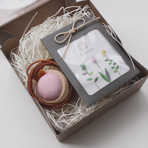 May include: A brown cardboard gift box filled with white paper shred containing a round wooden pincushion with a pink fabric top and a leather strap, alongside a dark gray card holder with a window displaying three floral-topped sewing pins.