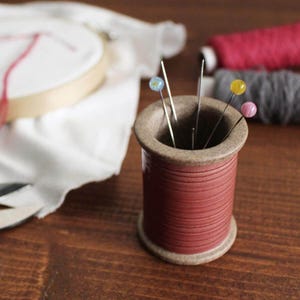 May include: A cylindrical ceramic pin cushion shaped like a vintage thread spool with a matte red body and textured beige rims. The top holds several sewing needles with colorful decorative heads in blue, yellow, and pink, standing upright.