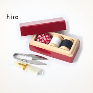 May include: A red wooden sewing box with the text hiro, containing a red and white floral pincushion, a white thread spool, and a black thread spool. Beside the box are silver metal thread snips and a small white fabric roll with gold trim.