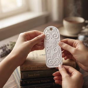 May include: A white, rectangular, lace-like bookmark with rounded top and bottom edges, featuring an intricate embossed floral and swirl pattern and a small circular hole at the top, held against a stack of classic hardcover books.
