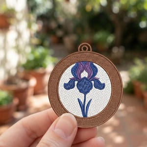 May include: A circular embroidered patch featuring a blue and purple iris flower centred on a white background, enclosed within a textured brown border with a small loop at the top for hanging or attachment.
