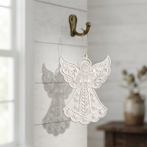 Può includere: Un ornamento decorativo a forma di angelo, bianco e piatto, caratterizzato da intricati motivi in rilievo sulle ali e sull'abito. L'angelo ha un'aureola sopra la testa ed è appeso con un filo sottile a un gancio da parete in metallo bronzato.