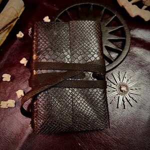 Puede incluir: Un diario rectangular de color marrón oscuro con un patrón texturizado de piel de serpiente en la cubierta. El diario se mantiene cerrado mediante una correa de cuero larga y fina de color marrón oscuro que se envuelve horizontalmente.