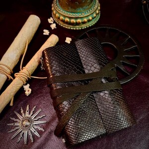 May include: A dark brown journal with a textured snakeskin pattern, secured by two crisscrossing leather straps. The journal is surrounded by two rolled parchment scrolls tied with twine, a metallic sun-shaped emblem, and a circular gear-like object.