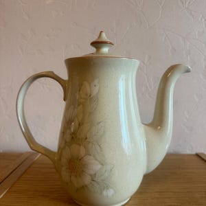 以下が含まれることがあります： 淡いクリーム色の陶磁器製ティーポット。斑点模様の仕上げが施され、白い花と緑の葉の繊細な花柄が描かれています。ポットには、尖ったつまみ付きの蓋、曲線的なハンドル、先細りの注ぎ口が付いています。