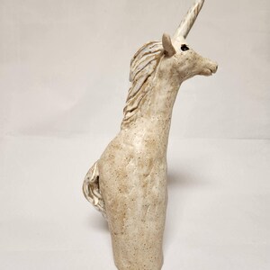 May include: A ceramic unicorn sculpture in a speckled, off-white colour. The mythical creature has a long, spiralled horn, a flowing mane, and a detailed tail. The sculpture is standing upright, with its head turned to the side.