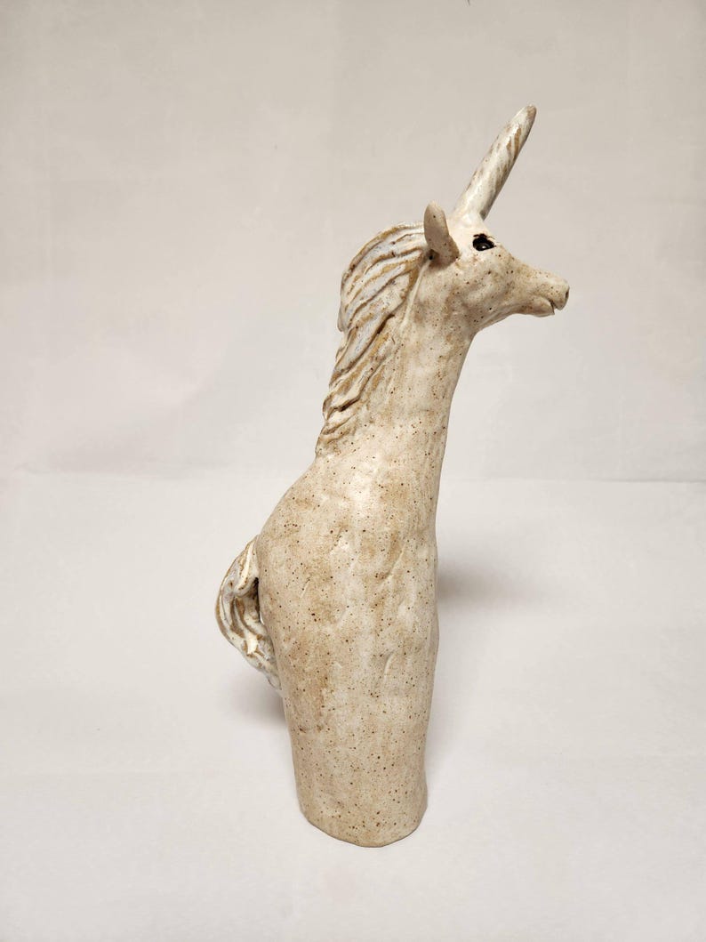 May include: A ceramic unicorn sculpture in a speckled, off-white colour. The mythical creature has a long, spiralled horn, a flowing mane, and a detailed tail. The sculpture is standing upright, with its head turned to the side.