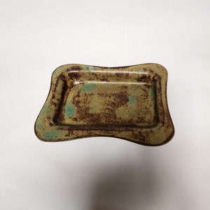 May include: A small, square ceramic dish with a wavy edge. The dish is glazed in a light brown color with green and brown speckles.