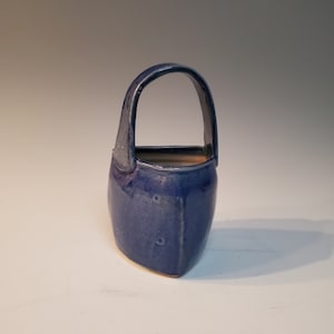 May include: A handmade, blue ceramic basket with a curved handle. The basket has a square base and a rounded top, with a glossy finish. The interior is a lighter, natural color. This decorative piece is suitable for home decor.