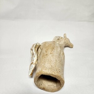 May include: A ceramic horse figurine in a light tan colour with a textured surface. The horse has a detailed mane and tail, with a hollowed-out body. The figurine is lying on its side.