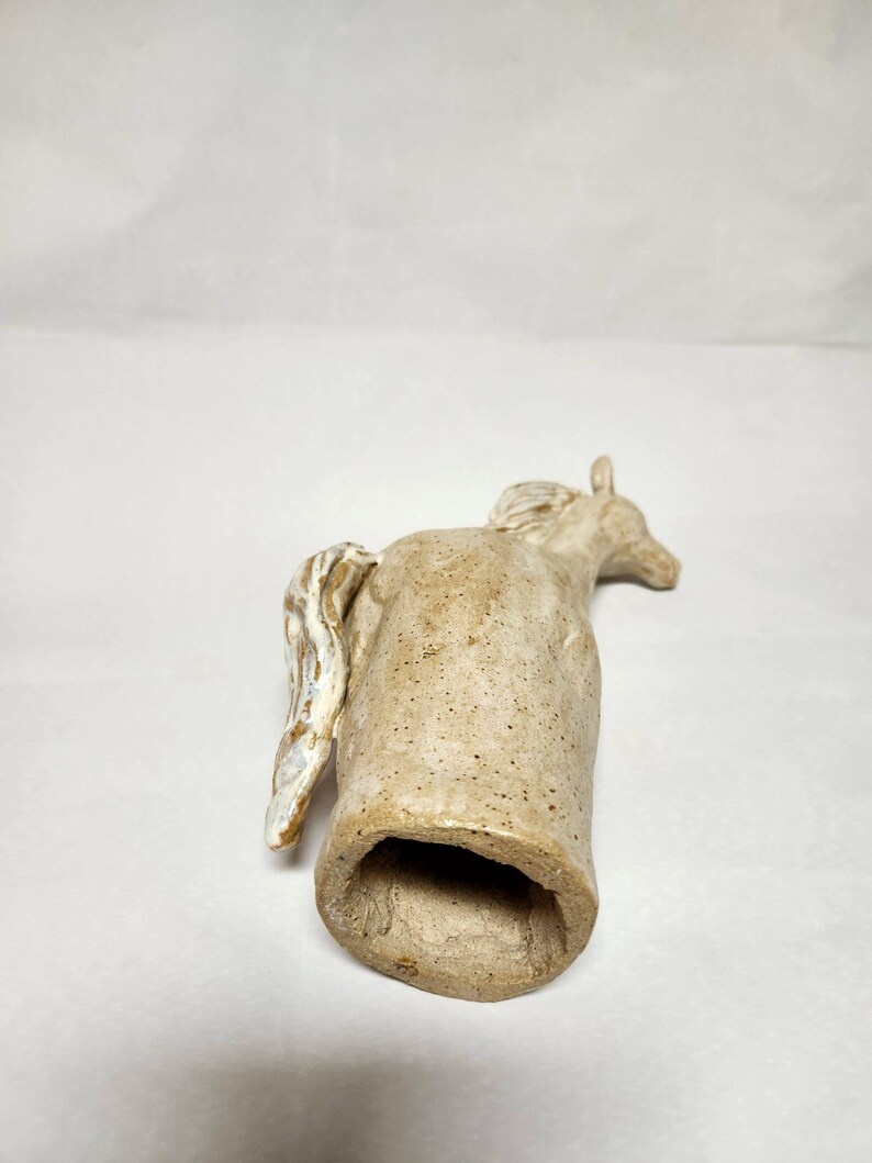 May include: A ceramic horse figurine in a light tan colour with a textured surface. The horse has a detailed mane and tail, with a hollowed-out body. The figurine is lying on its side.