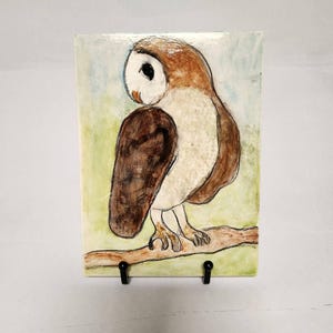 May include: A rectangular ceramic tile featuring a hand-painted owl perched on a branch. The owl has a white face, brown wings, and a light brown body. The background is a blend of green and blue hues, with a black stand.