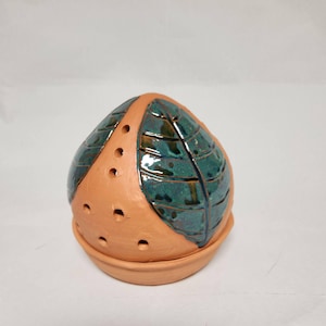 May include: A dome-shaped terracotta ceramic object with a circular base and a series of small ventilation holes. The surface features glossy, dark teal glazed leaf-shaped panels with etched vein details, contrasting with the matte clay finish.