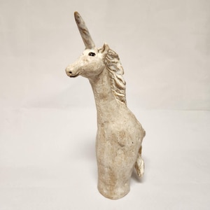 May include: A ceramic unicorn sculpture, off-white with brown speckles, featuring a long neck and a prominent horn. The mane is textured. The sculpture is a bust, with the body ending at the chest. A mythical creature.