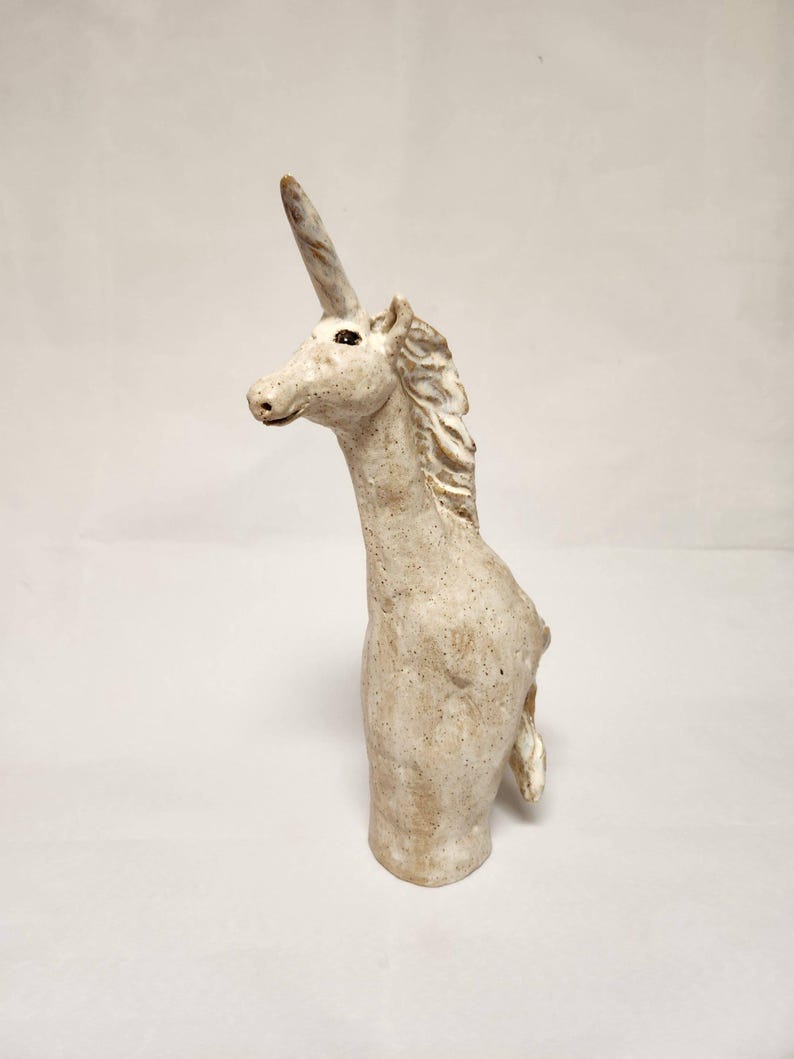 May include: A ceramic unicorn sculpture, off-white with brown speckles, featuring a long neck and a prominent horn. The mane is textured. The sculpture is a bust, with the body ending at the chest. A mythical creature.