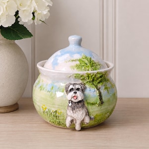 Puede incluir: Un tarro de cerámica con tapa que presenta un paisaje pintado a mano con un campo de hierba verde, un cielo azul con nubes y un árbol. Una figura tridimensional esculpida de un perro blanco y negro con la lengua rosa está fijada al frente.