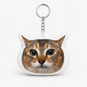 Op de afbeelding: Een pluche sleutelhanger met een realistische, hoge resolutie print van een bruin en tan cyperse kattengezicht. De kat heeft grote, expressieve groene ogen, een donkere neus en witte snorharen. Bevestigd aan een zilverkleurige metalen ring.