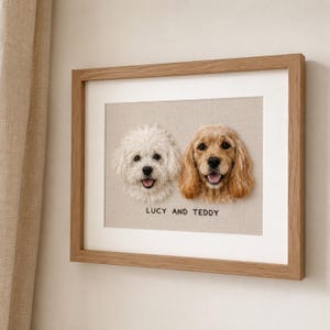 May include: A framed needle-felted portrait featuring a white curly-haired dog and a golden-brown spaniel-type dog side-by-side on a beige fabric background. Below the dogs, the text LUCY AND TEDDY is embroidered in black thread within a wooden frame.