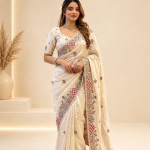 May include: An off-white saree featuring intricate floral embroidery in pink, blue, and green threads along the borders and pallu. The garment includes a matching blouse with short sleeves and a sweetheart neckline. The text PikanshStudio is visible.