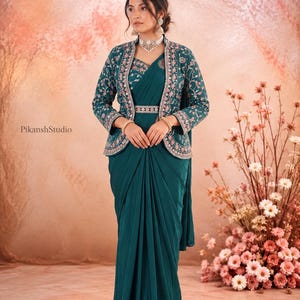 May include: A deep teal saree paired with a matching long-sleeved jacket featuring intricate pink and white floral embroidery along the edges. The ensemble includes a coordinating embroidered belt at the waist and a silver-toned choker necklace.