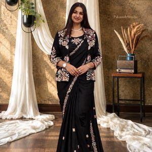 May include: A black saree paired with a matching black jacket featuring intricate multi-coloured floral embroidery. The saree has a decorative border with geometric patterns and small floral motifs, while the jacket has three-quarter sleeves.