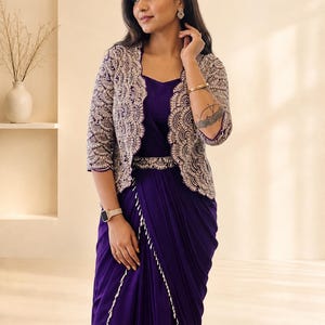 May include: A deep purple dress featuring a draped skirt with a white scalloped trim and a matching belt. The dress is paired with a cropped jacket that has three-quarter sleeves and an intricate silver and white scalloped embroidery pattern.
