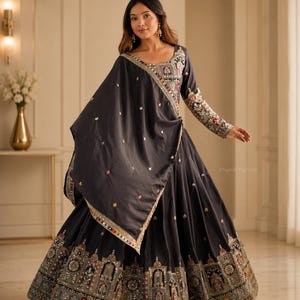 May include: A charcoal grey floor-length dress featuring long sleeves and a wide, flared skirt. The garment is adorned with intricate, multi-coloured embroidered panels and small mirror accents along the hemline, neckline, and sleeve cuffs.