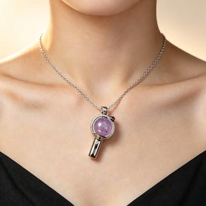 Puede incluir: Un collar de tono plateado con un colgante en forma de espejo de mano o varita. El colgante tiene un marco circular que sostiene una piedra preciosa redonda y facetada de color violeta claro, con una pequeña cuenta roja y un mango.