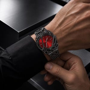 Könnte beinhalten: Eine Armbanduhr mit einem leuchtend roten, dreieckigen Zifferblatt, silbernen Zeigern und einer Datumsanzeige. Das Gehäuse und das Armband bestehen aus einem dunkelgrauen Material mit einem komplexen, netzartigen Gitterdesign.