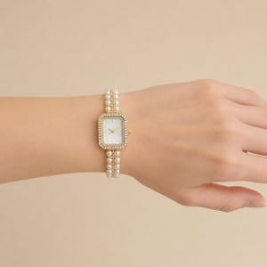 May include: A rectangular gold-tone wristwatch featuring a white face with gold hands. The watch case is bordered by small clear crystals, and the strap is composed of two parallel rows of white spherical faux pearls with gold-tone accents.