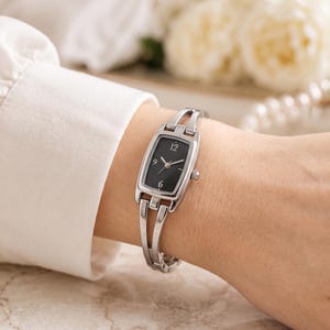 May include: A silver-toned analogue wristwatch featuring a rectangular black dial with white numerals at the 6, 9, and 12 o'clock positions. The watch has a slim, polished metal link bracelet with a split-band design connecting to the watch case.