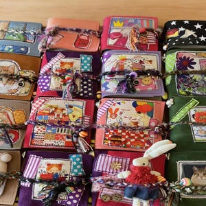 May include: A collection of small, rectangular fabric journals or booklets, each featuring unique patchwork designs, colorful illustrations, and decorative buttons. The items are tied with textured yarn and some include small plush animal attachments.