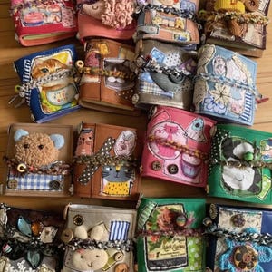 May include: A collection of sixteen small, rectangular fabric-covered journals, each featuring unique patchwork designs, decorative buttons, yarn ties, and various appliqués including animals, flowers, and patterns in a wide array of colors.