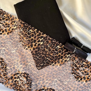 May include: A folded piece of black fabric, a wide strip of tan and black leopard print lace trim, and two small spools of black elastic trim. The leopard print lace features a repeating pattern of dark spots on a sheer, mesh-like background.