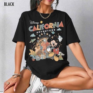 May include: A black short-sleeve t-shirt featuring a graphic print with the text Disney California Adventure. The design includes a colorful illustration of Mickey Mouse, a Ferris wheel, and various park elements in muted tones of orange, blue, and yellow.