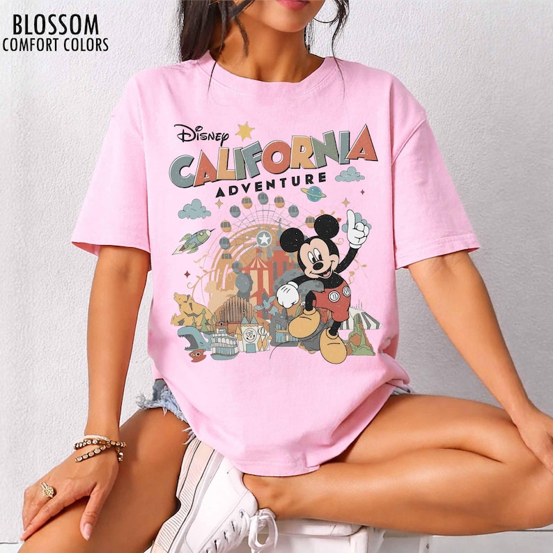 May include: A light pink short-sleeve t-shirt featuring a graphic print of Mickey Mouse and the text Disney California Adventure. The design includes a Ferris wheel, a rocket, and various park elements in muted tones. The text Blossom Comfort Colors appears in the top left corner.