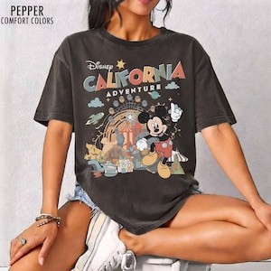 May include: A charcoal gray short-sleeve t-shirt featuring a graphic print with the text "Disney CALIFORNIA ADVENTURE". The design includes a cartoon mouse character, a Ferris wheel, and various park-themed icons in muted orange, blue, and cream colors.