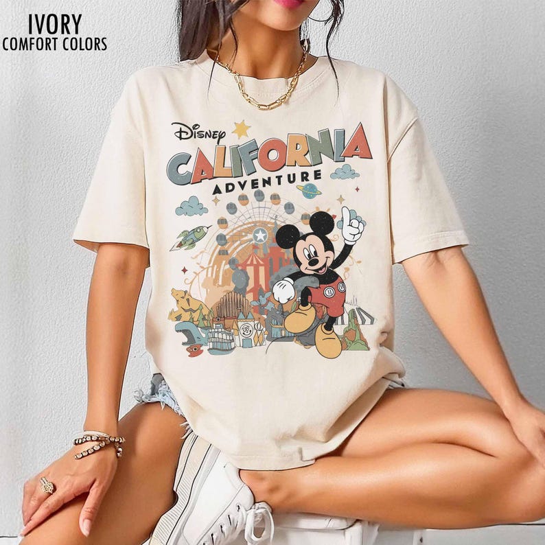 May include: An ivory short-sleeve t-shirt featuring a graphic print of Mickey Mouse and the text Disney California Adventure. The design includes a Ferris wheel, a rocket, and various park elements in muted tones. The shirt has a crew neckline.