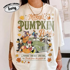 May include: An ivory t-shirt featuring a graphic print with the text Mickey's PUMPKIN Patch. The design includes cartoon characters, a green vintage truck, pumpkins, and autumn leaves. The text PUMPKINS - CORN MAZE - APHE CIDER Fresh from the patch is printed below the truck.