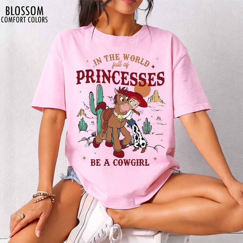 May include: A light pink short-sleeve t-shirt featuring a graphic of a cowgirl and a horse in a desert landscape. The text reads "IN THE WORLD full of PRINCESSES BE A COWGIRL". The brand name "BLOSSOM COMFORT COLORS" is printed in the top left corner.