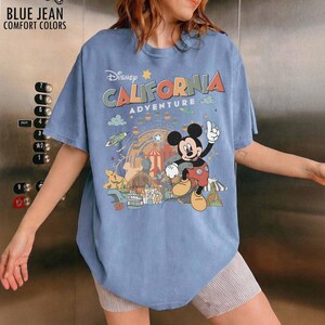 May include: A blue short-sleeve t-shirt featuring a graphic print of Mickey Mouse and the text Disney California Adventure. The design includes a Ferris wheel, park attractions, and small clouds. The shirt has a crew neckline and a relaxed fit.