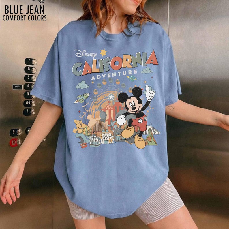May include: A blue short-sleeve t-shirt featuring a graphic print of Mickey Mouse and the text Disney California Adventure. The design includes a Ferris wheel, park attractions, and small clouds. The shirt has a crew neckline and a relaxed fit.