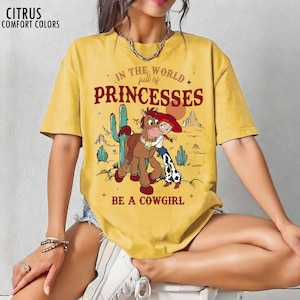 May include: A mustard yellow short-sleeve t-shirt featuring a graphic of a cowgirl and a horse in a desert landscape. The text reads: IN THE WORLD full of PRINCESSES BE A COWGIRL. The top left corner displays the text CITRUS COMFORT COLORS.