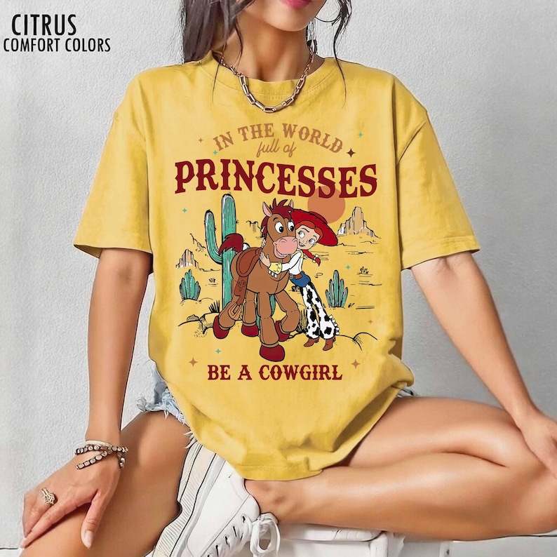 May include: A mustard yellow short-sleeve t-shirt featuring a graphic of a cowgirl and a horse in a desert landscape. The text reads: IN THE WORLD full of PRINCESSES BE A COWGIRL. The top left corner displays the text CITRUS COMFORT COLORS.