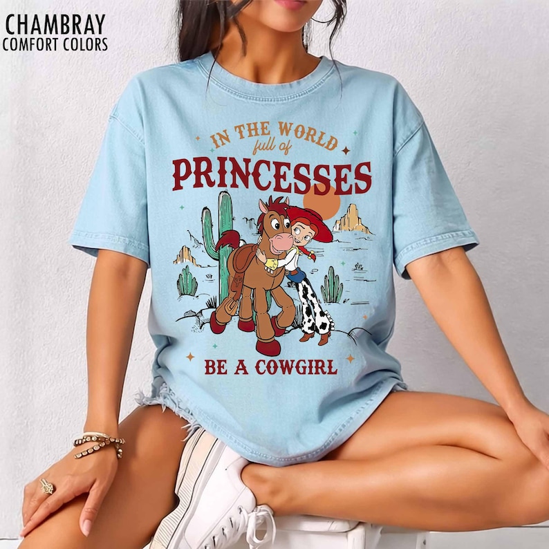 May include: A light blue t-shirt featuring a graphic of a cowgirl hugging a horse in a desert landscape with cacti and mountains. The text reads "IN THE WORLD full of PRINCESSES BE A COWGIRL" in red and brown lettering. The shirt is labeled "CHAMBRAY".