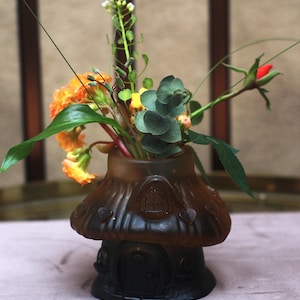Puede incluir: Un jarrón de vidrio esmerilado marrón con forma de casa de hongo con un sombrero ancho y una puerta central, lleno de un ramo de flores naranjas, hojas de eucalipto verdes y un capullo de rosa roja, creando un arreglo floral decorativo.