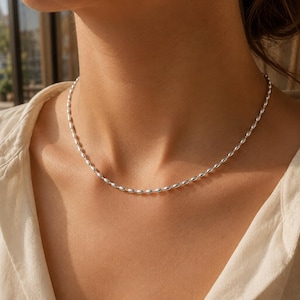 Op de afbeelding: Een zilverkleurige ketting bestaande uit een doorlopende reeks kleine, langwerpige, rijstvormige kralen. De metalen schakels hebben een glad, reflecterend oppervlak dat het licht opvangt, wat zorgt voor een uniforme en verfijnde uitstraling.