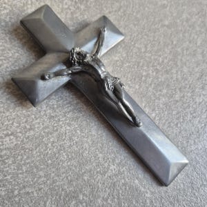 May include: A metallic gray crucifix featuring a smooth, beveled cross and a detailed three-dimensional figure of Jesus Christ attached to the center. The object has a matte finish and a clean, angular design with pointed ends on the cross arms.